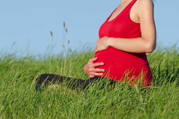 Pregnant woman in red top cradling belly while sitting in grass outdoors