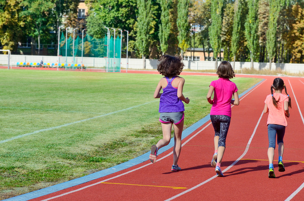 Three children running on a red athletic track at a sports field with green grass and trees in background