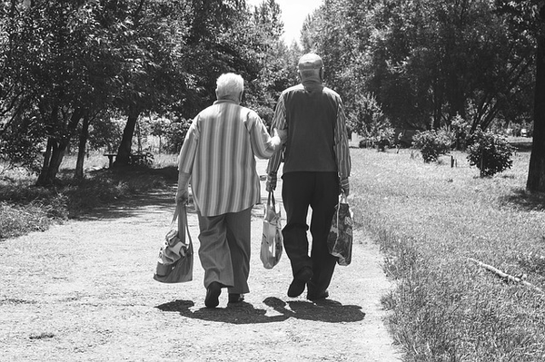 Two elderly people walking together on a tree-lined park path holding hands