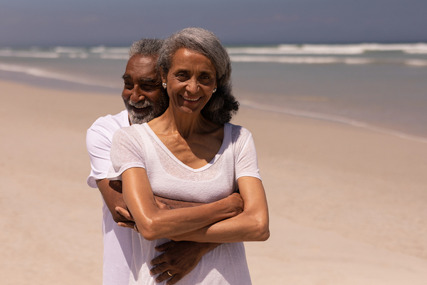 Two senior adults smiling at the beach, man behind woman with arms around her shoulders