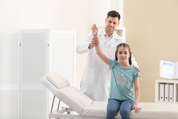 Male chiropractor in white coat giving high-five to young girl in medical office with examination table
