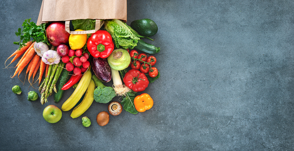 Fresh vegetables and fruits spilling from a paper grocery bag on a dark surface, including carrots, lettuce, peppers, tomatoes, cucumbers, bananas, and apples.