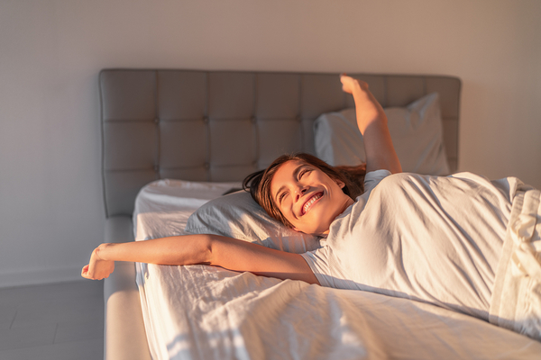 Person lying in bed with arm stretched out, smiling in morning sunlight