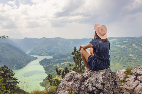 Woman sitting on rocky outcrop wearing hat, overlooking mountain valley with turquoise river below