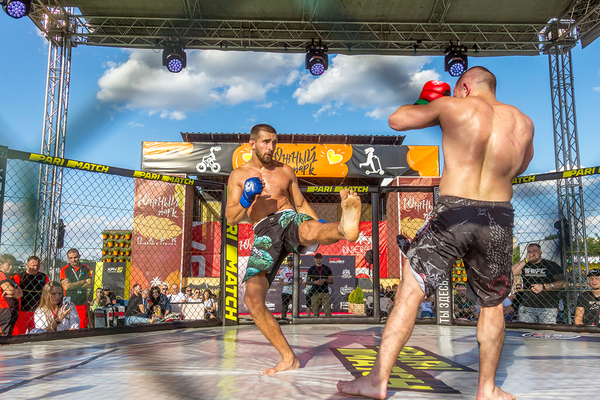 Two shirtless men boxing in a ring under a metal truss structure with spectators and banners visible.