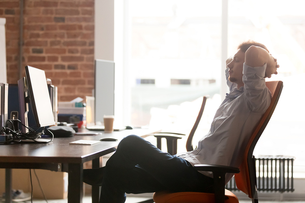 Man reclined in office chair at desk with computer, hands behind head in relaxed pose