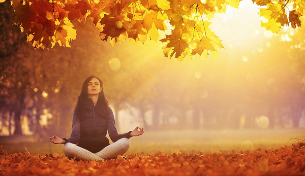 Woman meditating in lotus position under golden autumn foliage surrounded by fall leaves