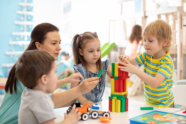 Caregiver supervising children building with colorful wooden blocks and toy vehicles at a table
