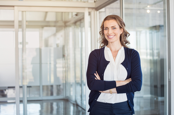 Professional woman in navy cardigan and white blouse standing with arms crossed in modern office with glass walls