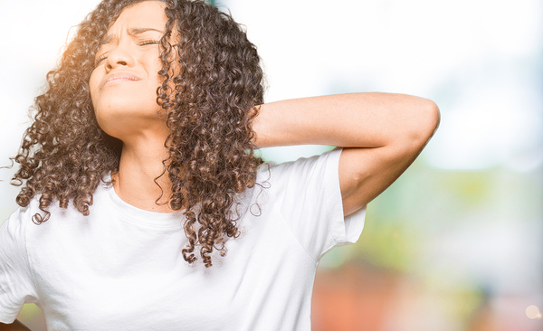 Woman with curly brown hair touching her neck, wearing white t-shirt outdoors