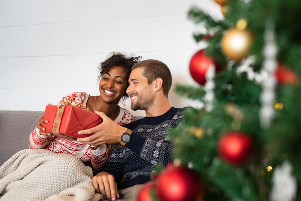 Smiling couple sitting on couch holding a red wrapped gift with Christmas tree and ornaments in foreground