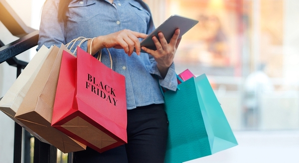 Person in denim shirt holding tablet while carrying colorful shopping bags labeled Black Friday