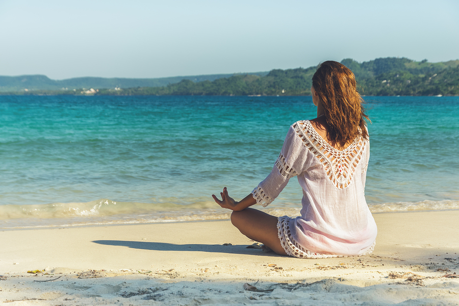 Woman in white crocheted cover-up sitting in yoga pose on sandy beach overlooking turquoise ocean and forested coastline
