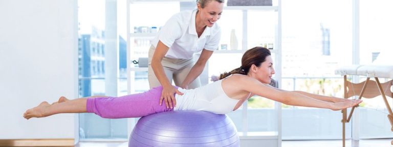 Woman balancing on purple exercise ball with trainer providing instruction in bright fitness studio with city skyline view