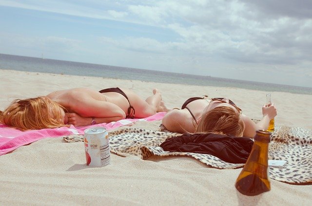 Two people lying on beach towels in the sand near the ocean with beverages and sunscreen.