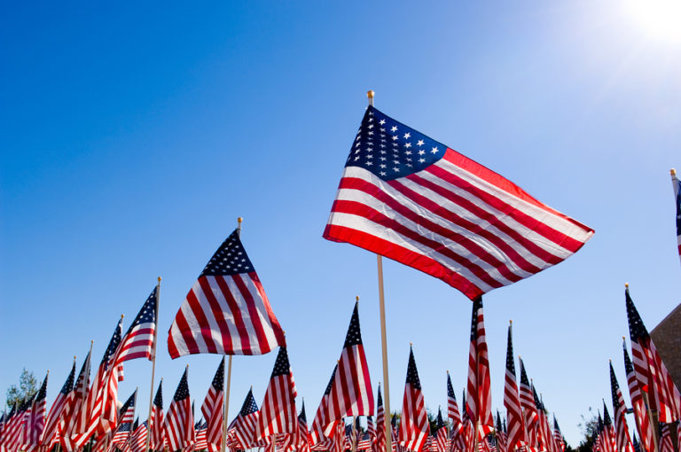Multiple American flags displayed against a blue sky, with one larger flag prominently featured in the center.