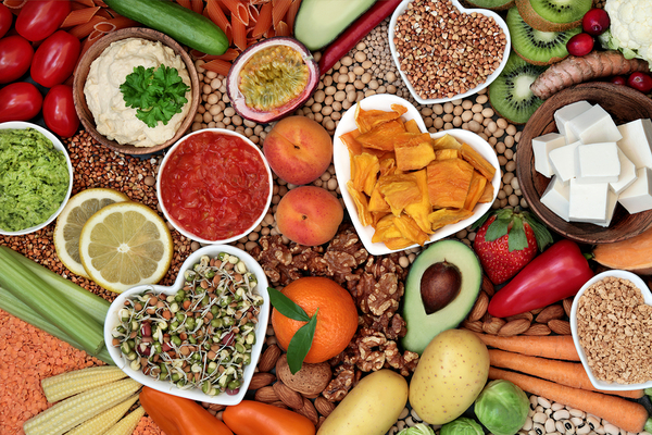Overhead view of healthy foods including vegetables, fruits, legumes, nuts, seeds, and plant-based proteins arranged on a neutral surface.