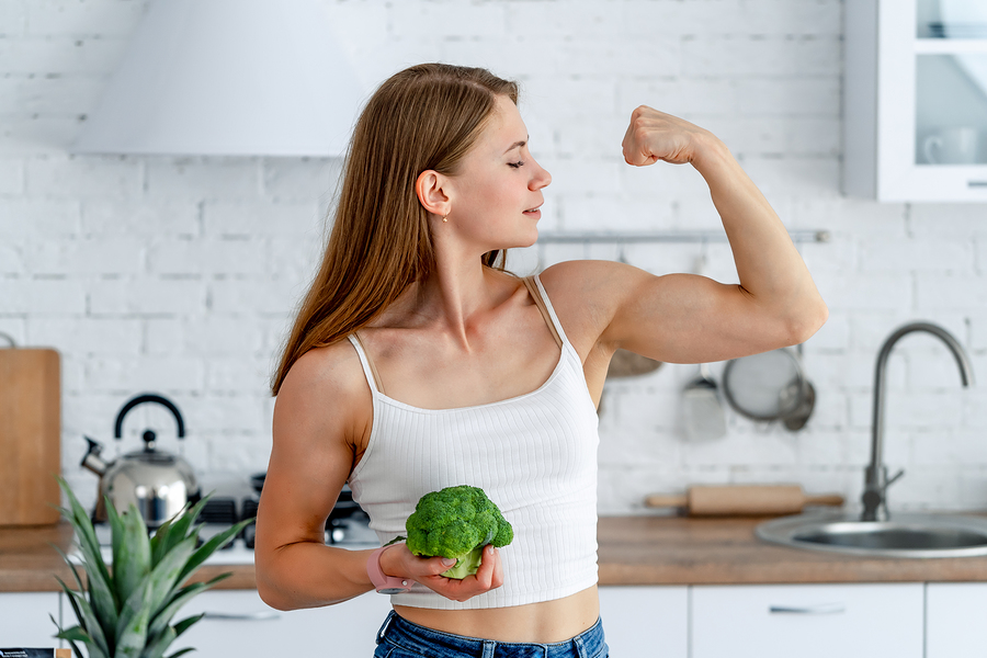 Woman in kitchen flexing bicep while holding fresh broccoli