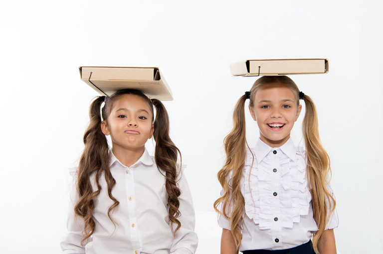 Two smiling schoolgirls wearing white shirts with cardboard boxes balanced on their heads against a white background.