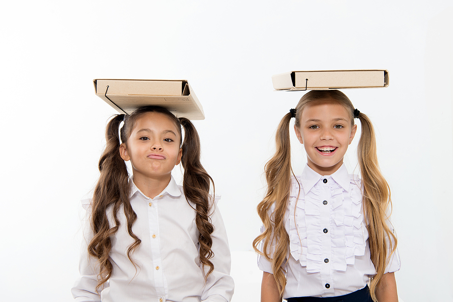 Two smiling schoolgirls wearing white shirts with cardboard boxes balanced on their heads against a white background.