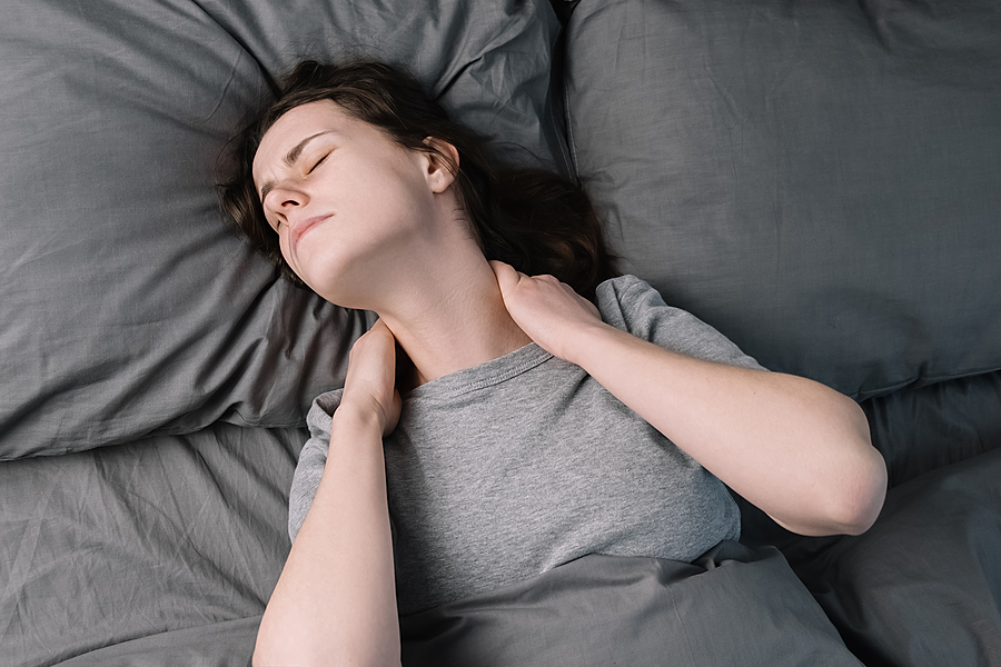 Woman lying on gray bedding with hand on neck, eyes closed in discomfort