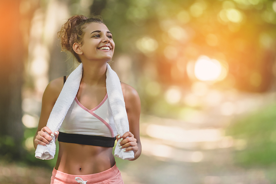 Woman in athletic wear with towel around neck smiling outdoors on a tree-lined path