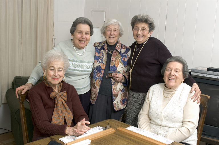 Five older women smiling at a table indoors, some standing and two seated