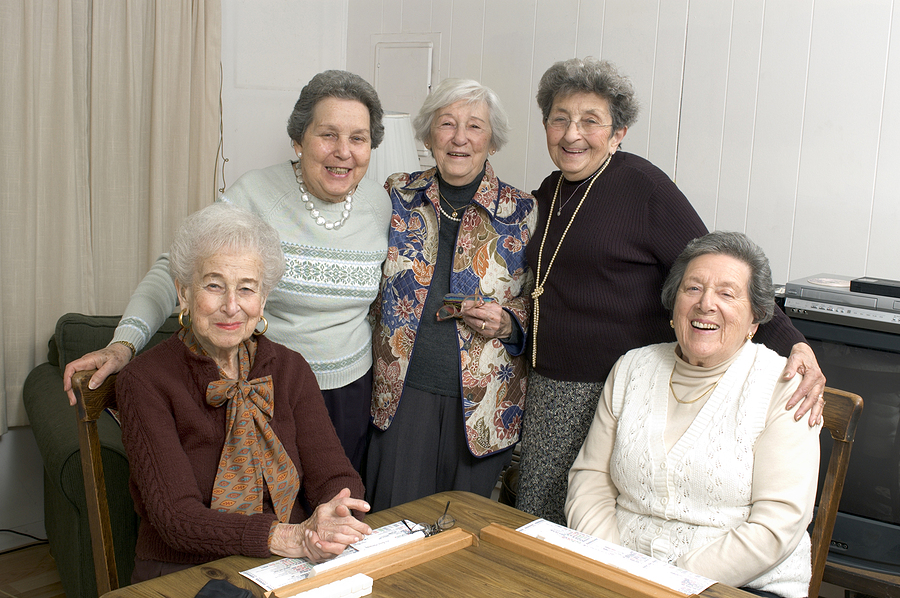 Five older women smiling at a table indoors, some standing and two seated