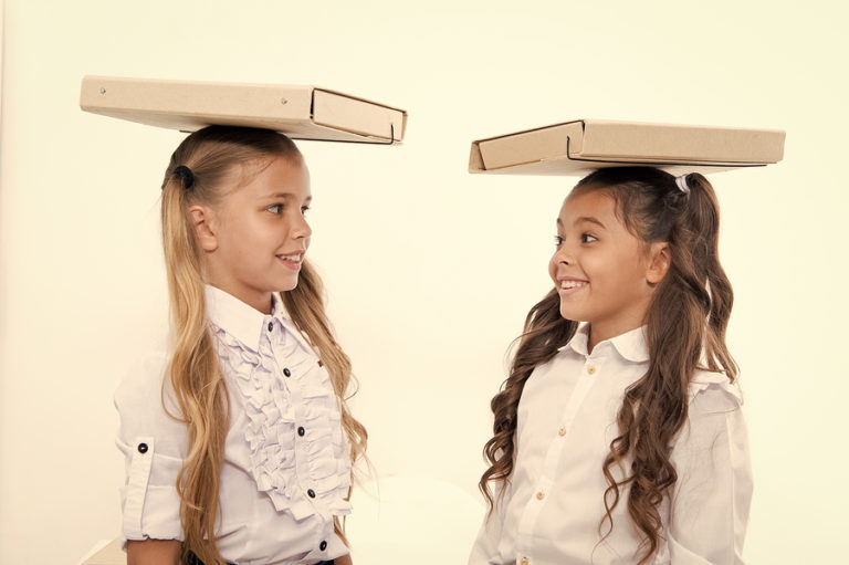 Two schoolgirls balancing cardboard boxes on their heads while looking at each other smiling