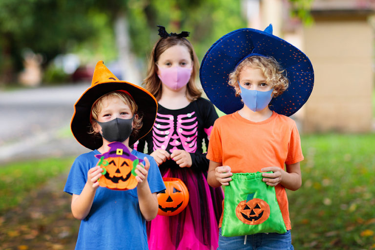 Three children wearing Halloween costumes and masks hold jack-o'-lantern candy buckets while trick-or-treating outdoors