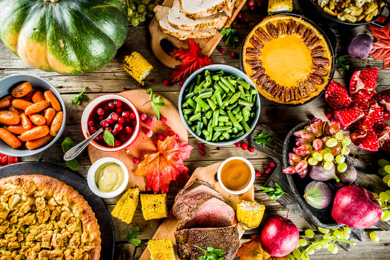 Overhead view of fall harvest foods including roasted vegetables, fresh berries, corn, squash, bread, and prepared dishes on wooden table