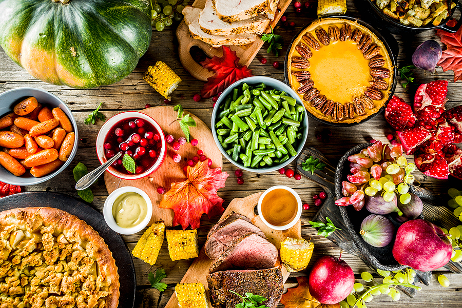 Overhead view of fall harvest foods including roasted vegetables, fresh berries, corn, squash, bread, and prepared dishes on wooden table