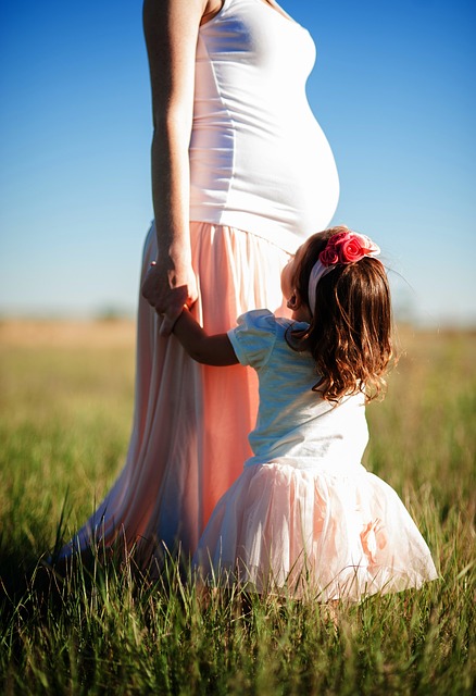 Pregnant woman in white and coral dress standing in grassy field with young child in flower crown holding her skirt