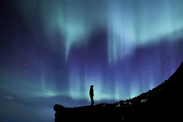 Silhouetted figure standing on rocky outcrop beneath vibrant aurora borealis with blue and green lights dancing across starry night sky