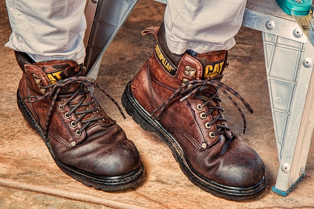 Person wearing worn brown leather work boots with white socks and light-colored pants standing on wooden floor