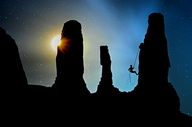 Silhouette of rock formations at night with a climber rappelling down and a glowing moon or light source behind the peaks
