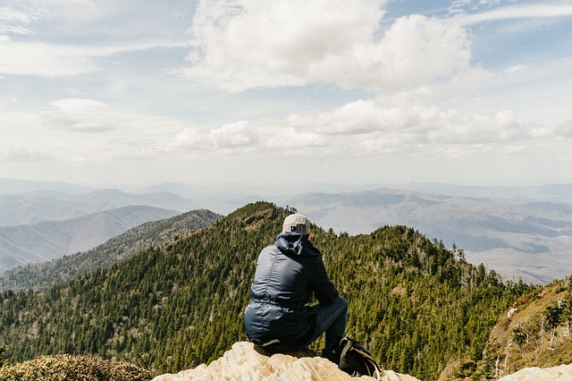 Person in blue jacket and white cap crouched on rocky outcrop overlooking forested mountain ranges and valleys
