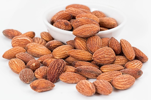 Raw almonds scattered on white surface with bowl of almonds in background