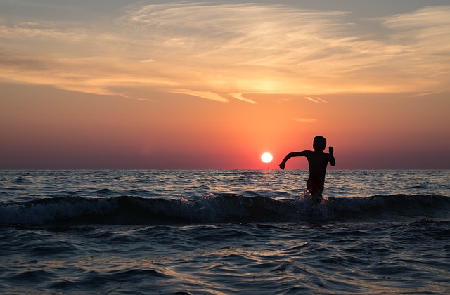 Surfer in wetsuit holding surfboard wading into ocean at sunset with golden sky