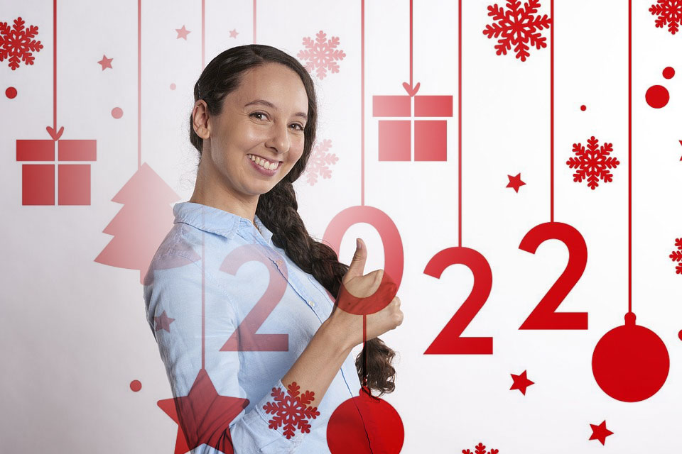 Girl smiling at camera with long dark hair wearing light blue shirt, standing in front of festive Christmas backdrop with hanging ornaments, snowflakes, and gift decorations