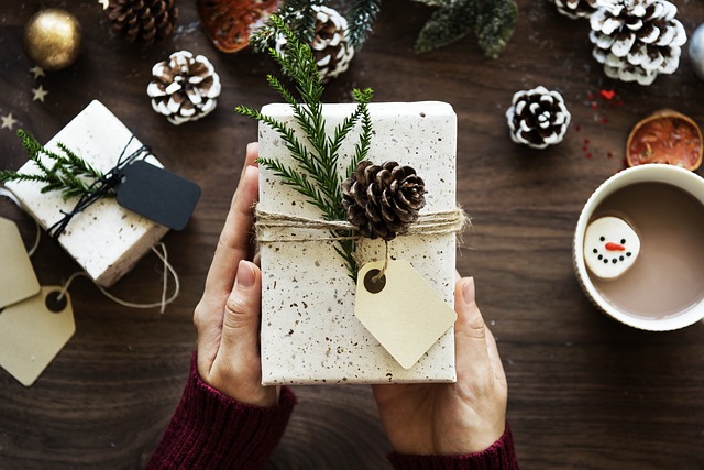 Hands holding a wrapped gift decorated with greenery and pinecone on wooden surface surrounded by holiday decorations