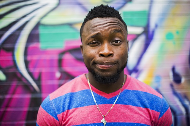 Man with beard and styled black hair wearing colorful striped shirt and cross necklace, standing in front of vibrant graffiti wall