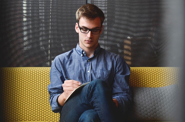Young man wearing glasses and denim shirt sitting on yellow bench writing in notebook