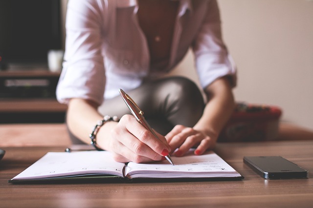 Person writing in open notebook with pen at wooden desk
