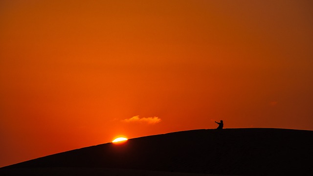 Person silhouetted on hilltop against orange sunset sky with sun on horizon