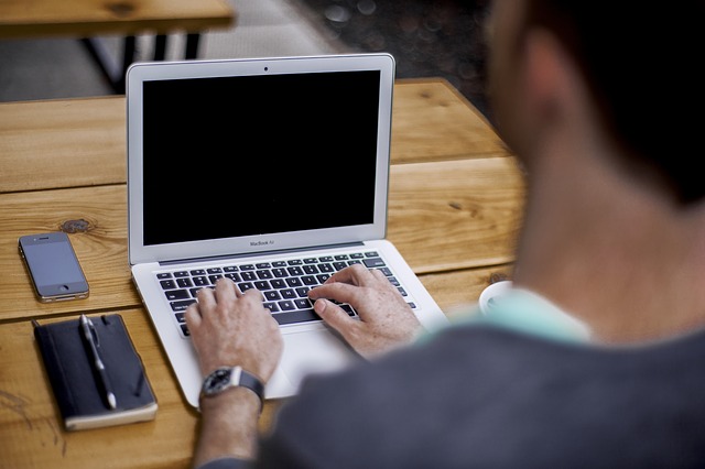Person typing on laptop at wooden table with smartphone nearby