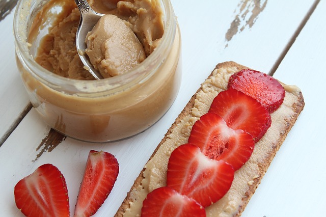 Peanut butter in a jar with spoon, strawberry slices on whole grain toast, and loose strawberry pieces on white wooden surface