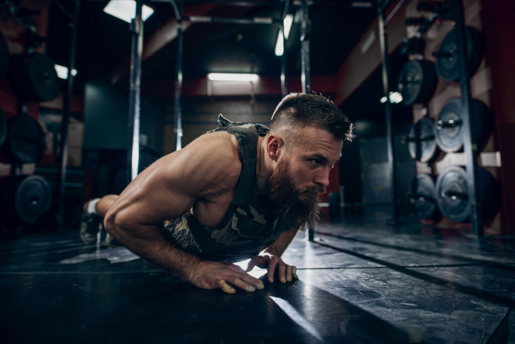 Bearded man in athletic tank top performing a plank exercise on dark gym floor with weights visible in background