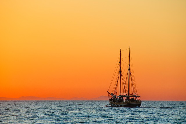 Silhouetted sailing ship with two masts on calm ocean water at sunset with golden orange sky