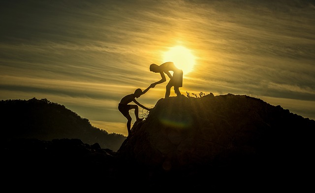 Silhouette of one person helping another climb up a rocky hill against a bright sunset sky